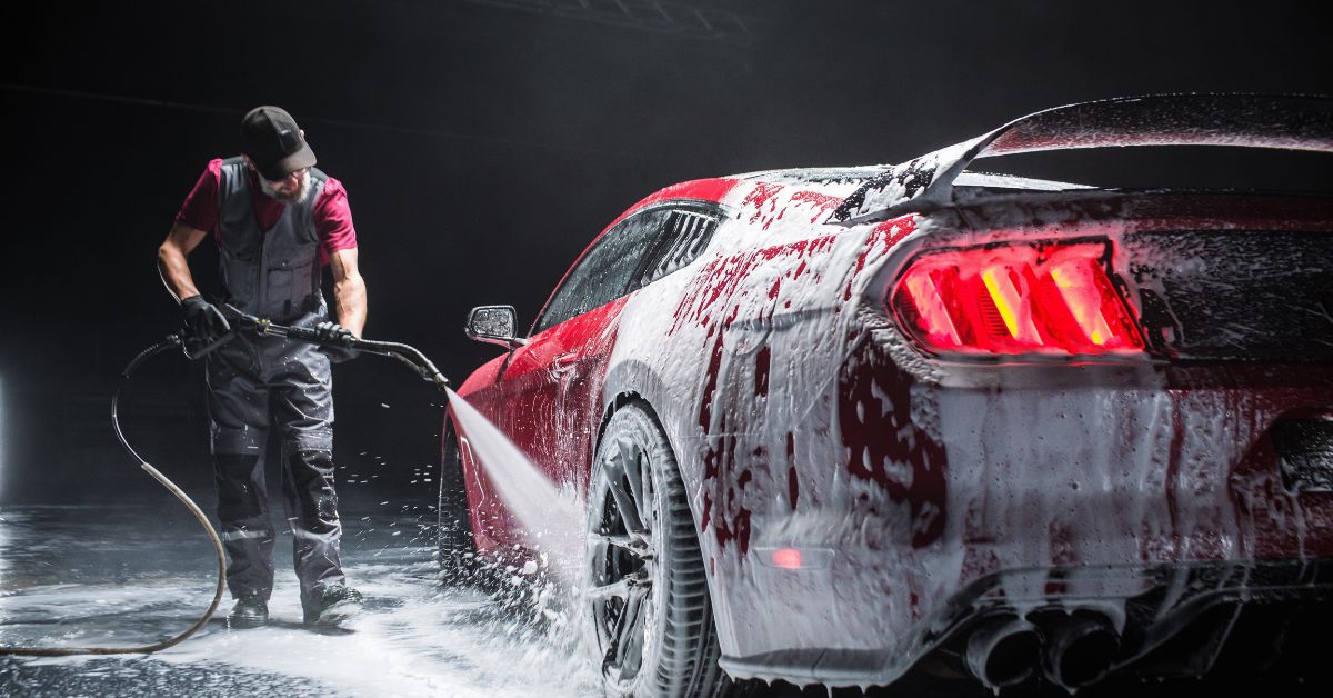 Automotive Detailer Washing Away Smart Soap and Foam with a Water High Pressure Washer. Close Up of a Red Performance Car Getting Care and Treatment at a Professional Vehicle Detailing Shop